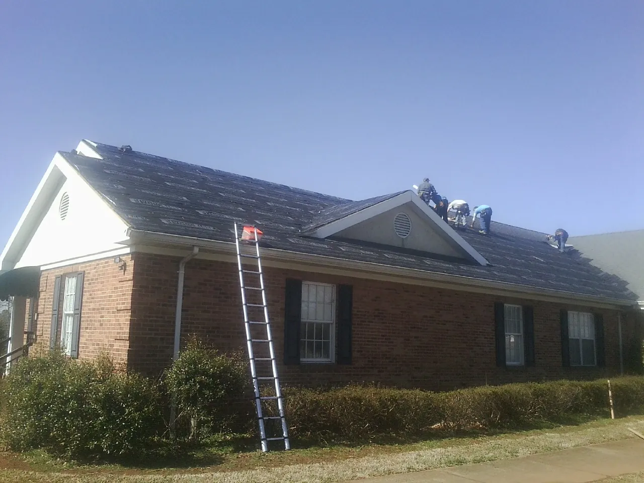 Crew tearing shingles off a brick ranch roof with a ladder set — Metal Roof Repair work by our crew in Amherst